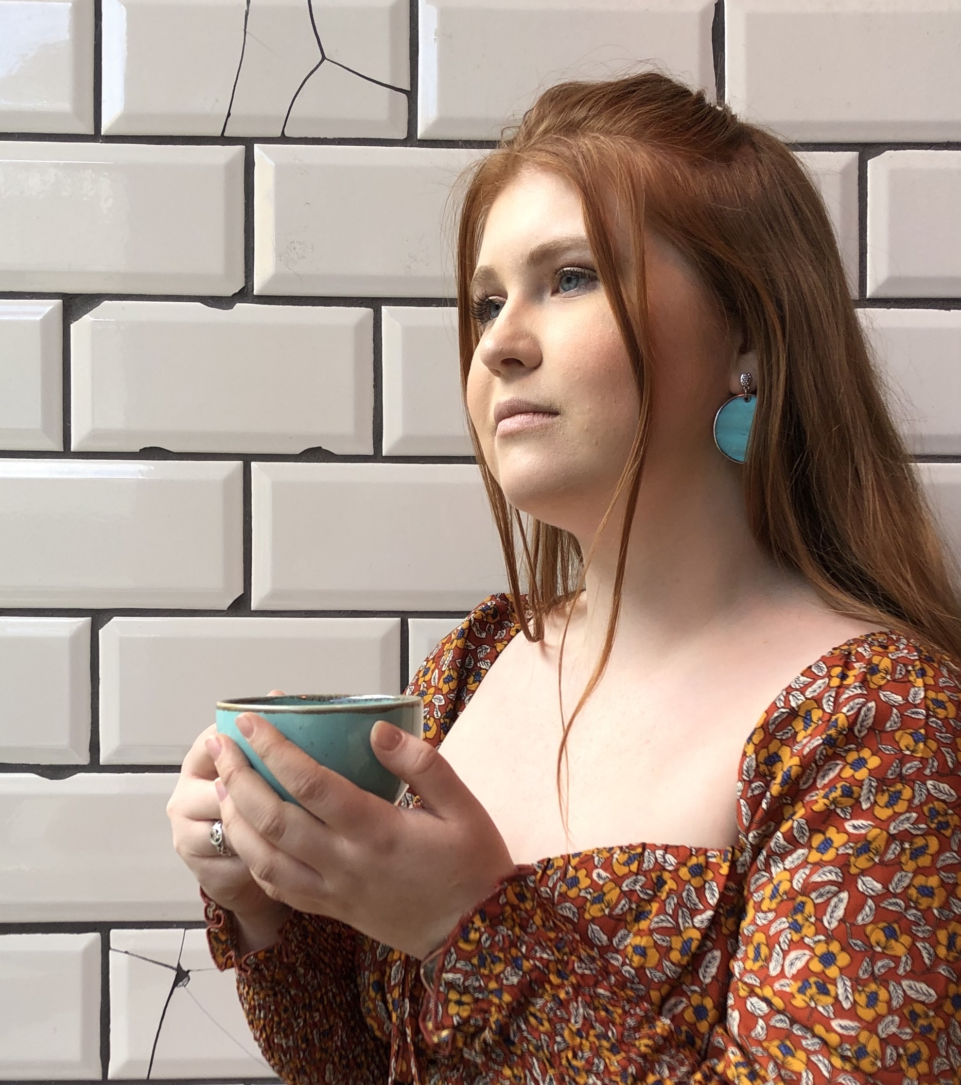 Charlotte is pictured sitting next to distressed white tiles. She holds a turquoise coffee cup which matches her earrings and looks off camera.